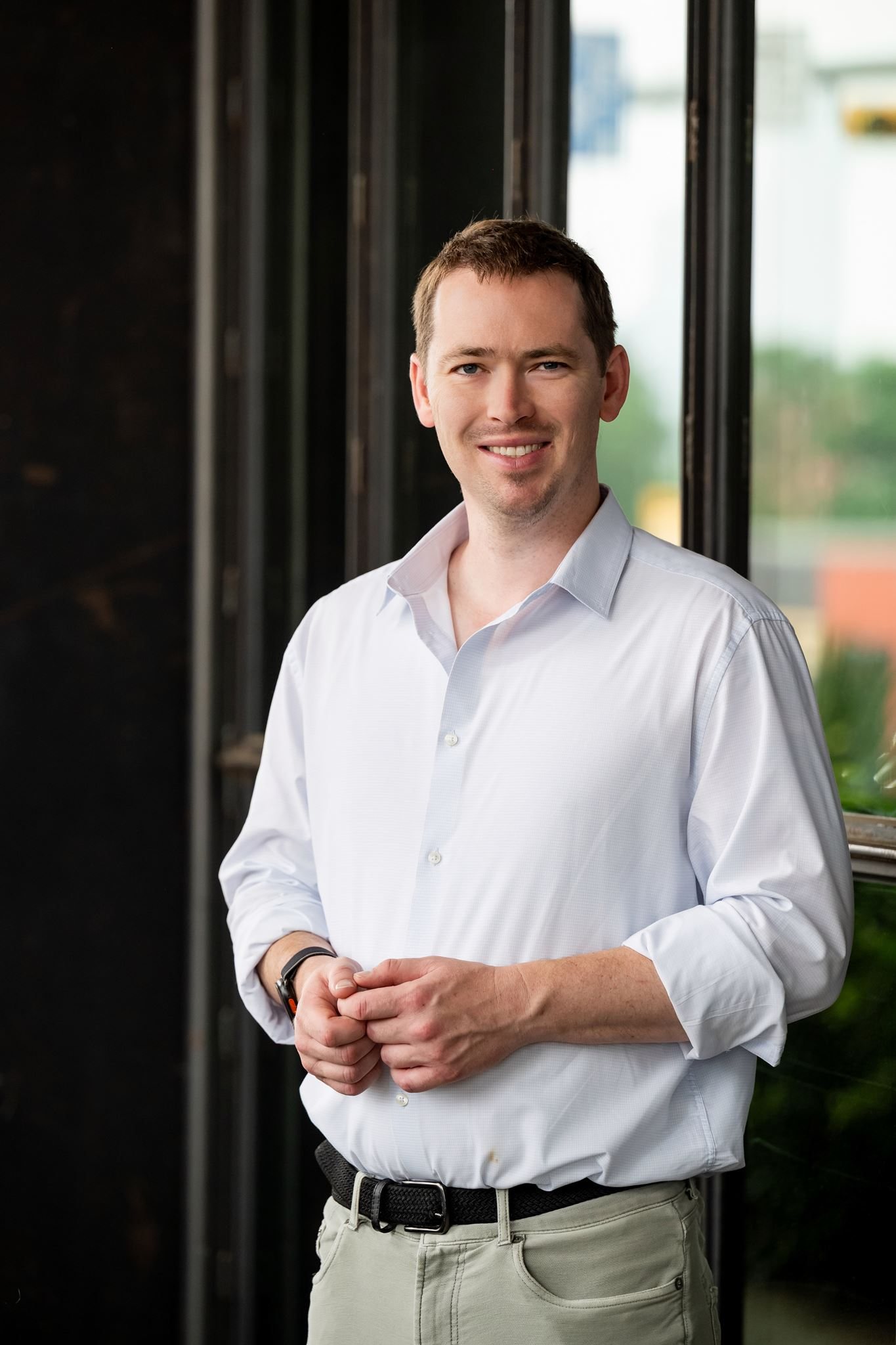 Jon Lofquist in a white shirt, standing relaxed by a city window