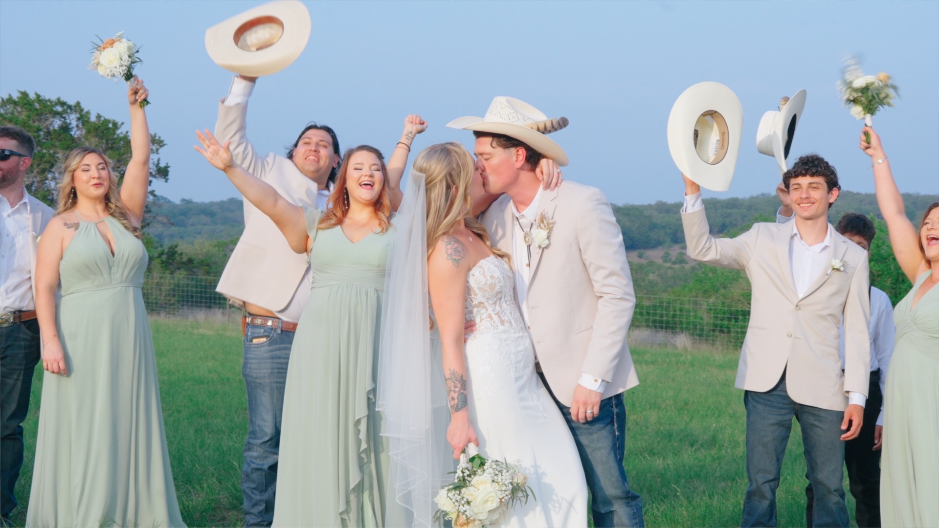 Wedding party cheering and throwing their hats as the couple kiss in an open Texas field at dusk