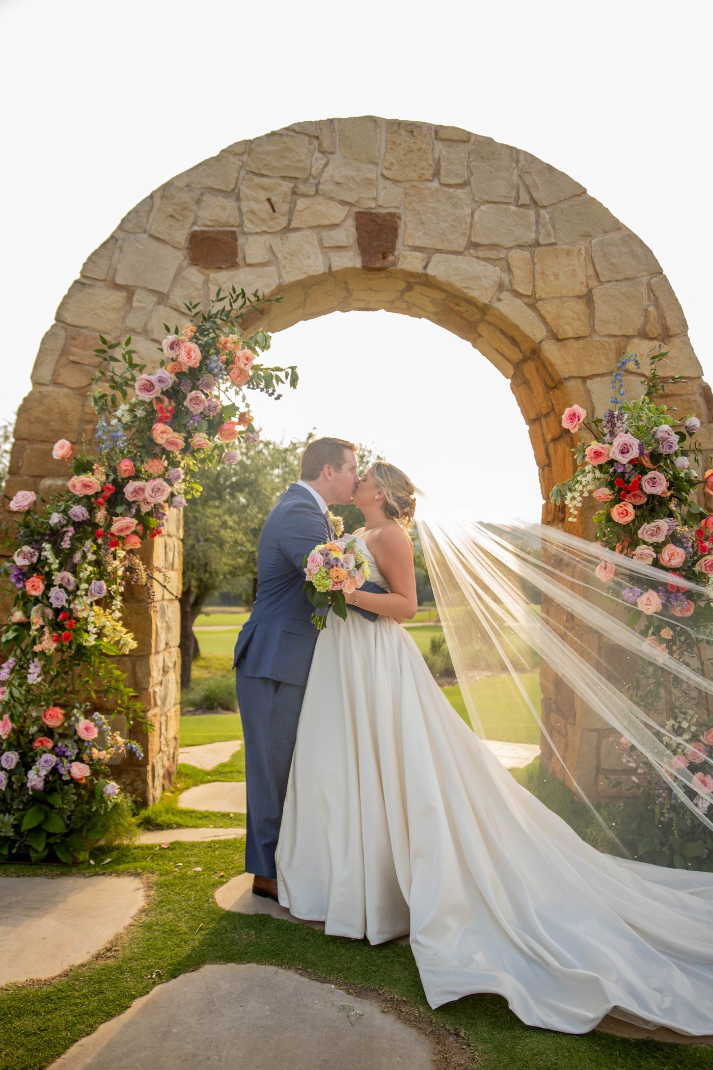 A bride and groom beneath a stone arch, her veil catching the golden sunlight